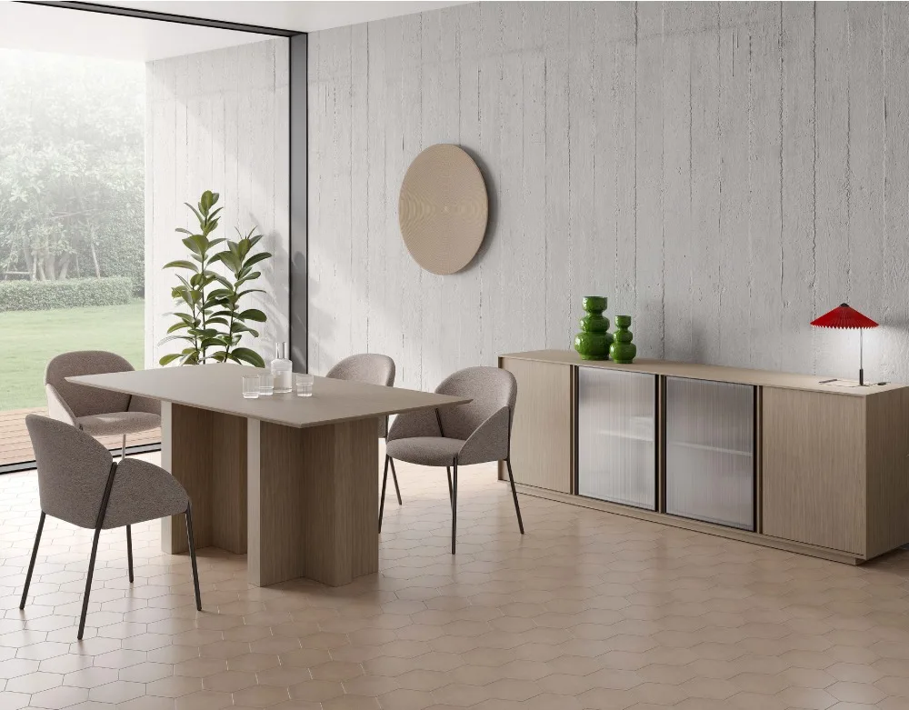 Fotografía de un comedor moderno en tonos tostado y gris claro. A la izquierda , la mesa de comedor de madera y cuatro sillas de tela tostada a su alrededor. Detrás, una planta de suelo alta y un gran ventanal que ocupa toda la pared y da a un jardín arbolado. A la derecha, en la pared, hay un aparador de la misma madera que la mesa de comedor, con las dos puertas centrales de cristal ahumado. Sobre el aparador, dos jarrones verdes a la izquierda y una lámpara de mesa roja a la derecha. La pared es de hormigón, mientras que el suelo es de azulejo tostado hexagonal.